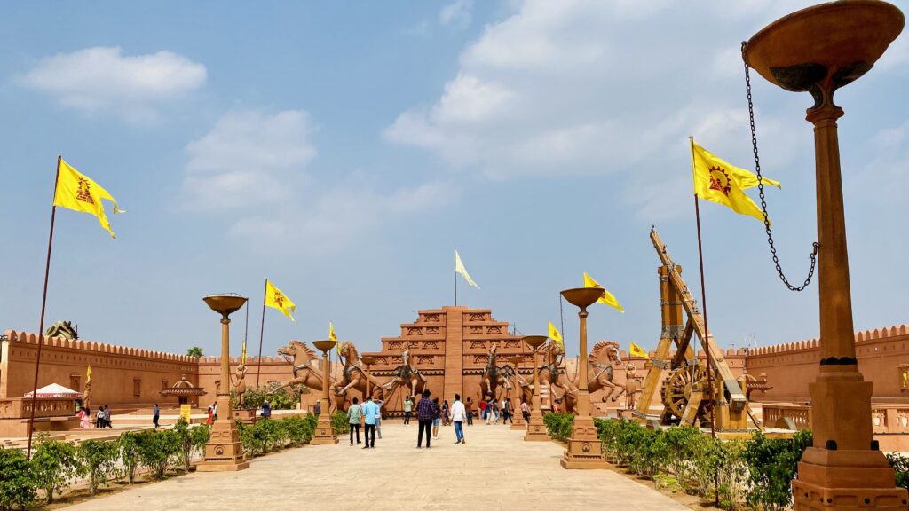 Wide front courtyard of the Mahishmati kingdom set from Baahubali at Ramoji Film City. A broad walkway leads to a central staircase with a throne at the top. Tall royal flags wave on both sides, and rows of large horse statues stand along the square. Visitors are walking through the space under an open sky.